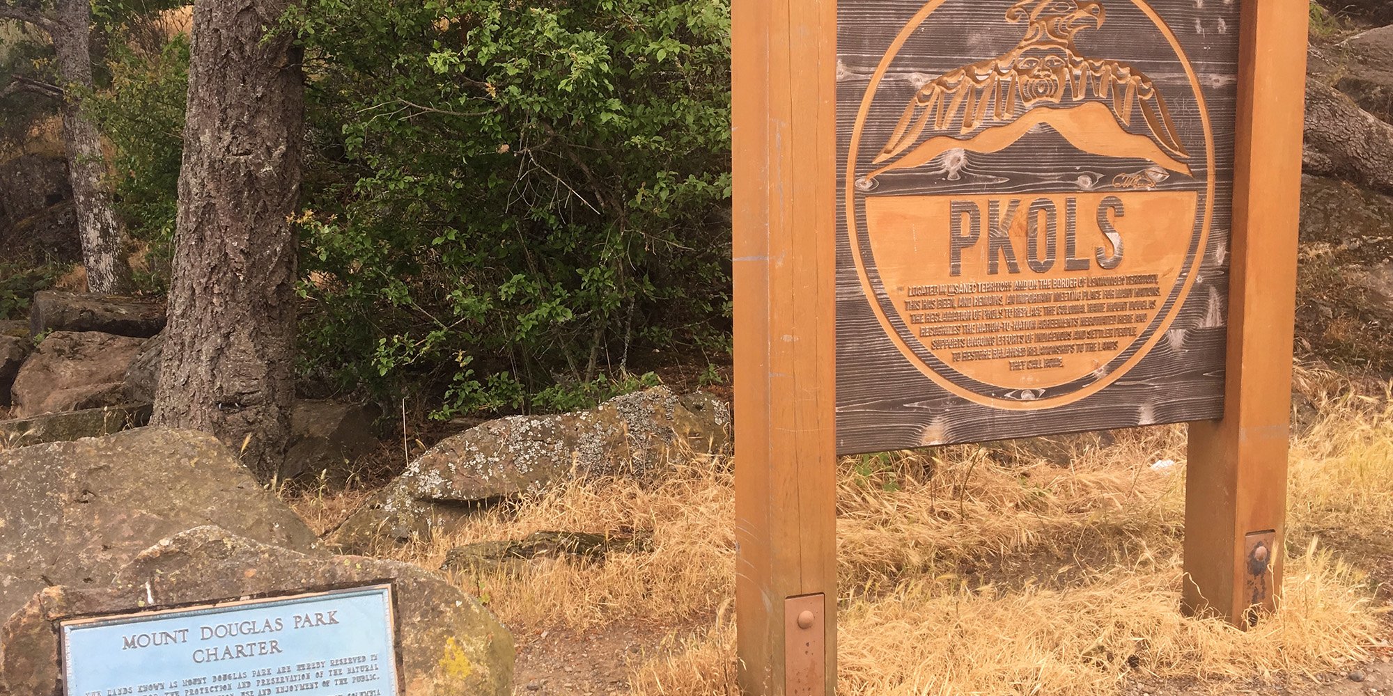 Two signs at the top of Mt. Doug/Pkols in Victoria, BC; one comes from the Canadian government, who refer to the mountain as Mt. Doug, while the other, newer, sign refers to the mountains traditional name, Pkols.